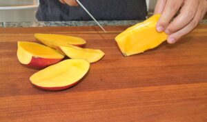 two hands with a large knife slicing mangos on a mahogany butcher block cutting board 