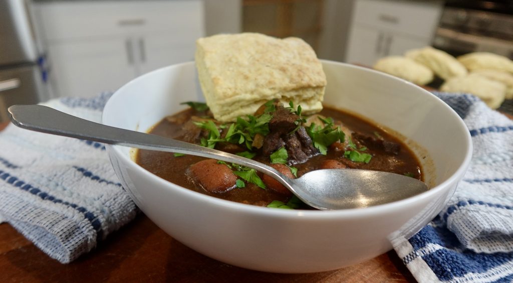 Beef stew on cutting board in a bowl with a biscuit and spoon. 